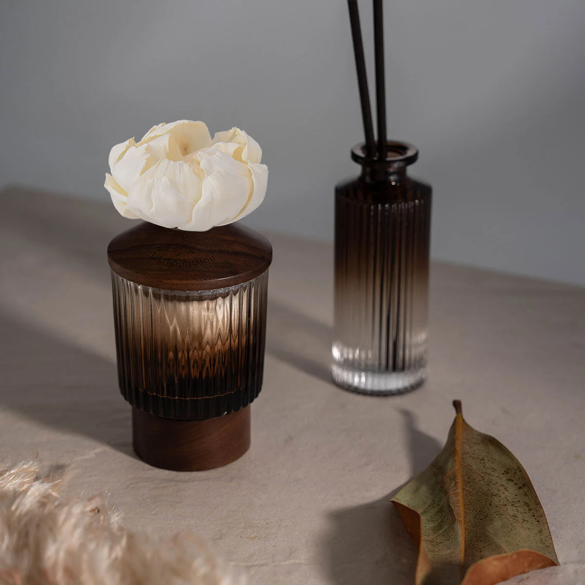 Two LiliaMansion reed diffusers in ribbed smoked glass with walnut lids, one topped with a white sola flower, on a warm stone surface